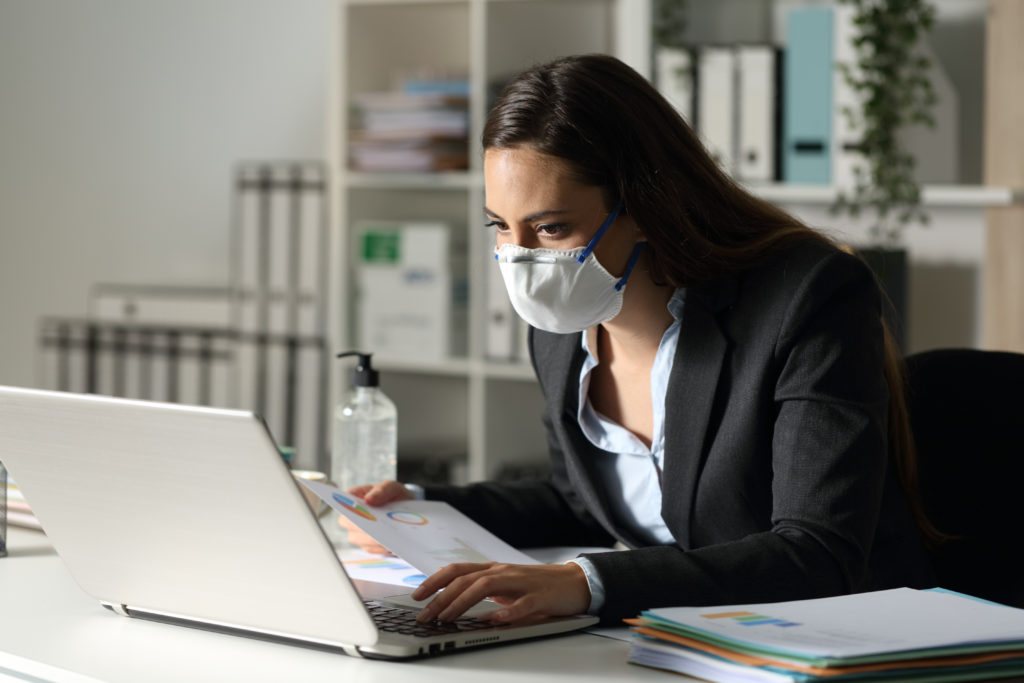 A woman wearing a mask works alone on a laptop with a bottle of hand sanitizer visible in the background